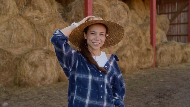 Portrait of cheerful young woman wearing straw hat and plaid blue shirt smiling at camera while standing in barnyard against hay bales stacked under shed, copy space