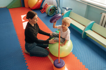 On the ball, putting rounded toys on the stick, balance. Preschool child with teacher during developmental activities in a playroom