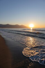 A stunning sunset over the ocean casts a golden reflection on the beach, with waves gently rolling to the coast under a vibrant, cloud-filled dusk sky