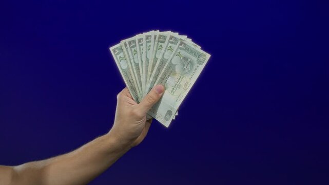 Caucasian man holds uae dirham banknotes against a blue background, showcasing currency with an isolated hand gesture.