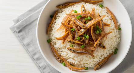 Bowl of rice topped with fried anchovies and green onion garnish, displayed on a gray cloth and white wood background, studio shot, overhead view.