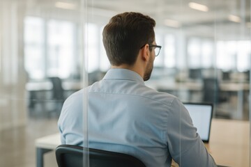 Professional Working in Modern Office. Rear view of a man with glasses working on a laptop at his desk in a modern, open-plan office.