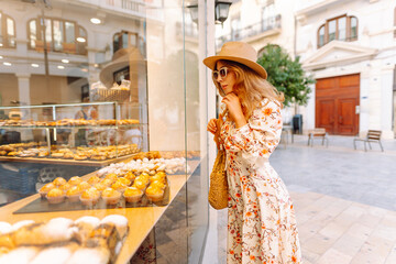 A young woman in a floral dress and wide-brimmed hat stands by a bakery window, looking at delicious pastries and holding a drink. The sun shines brightly on the charming street. © Koval988