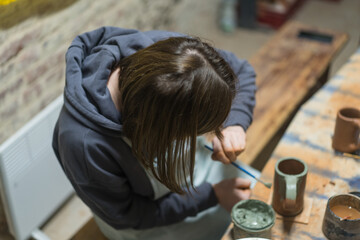 Girl paints her own handmade clay mug. Pottery teaching lesson. Potter makes dishes from clay, ceramics. Top view.