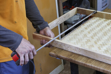 Focused craftsman measuring handmade wooden frame with woven bamboo bottom in workshop. This carpenter uses tape measure for precise DIY furniture project