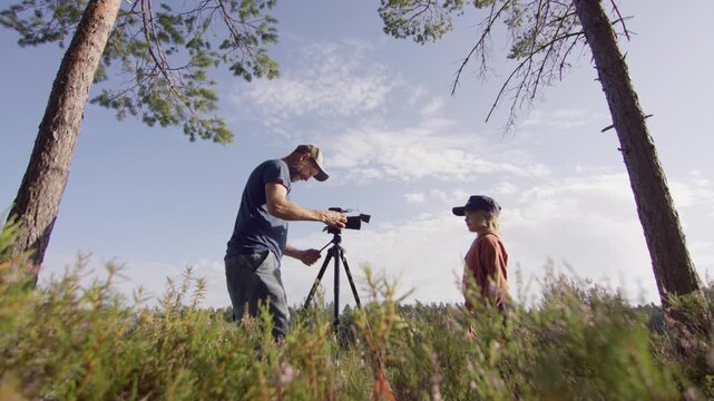Videographer use camera on tripod in woods to record video of young boy, profile