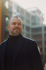 Fototapeta premium Portrait of positive elegant man in coat looking away on urban street