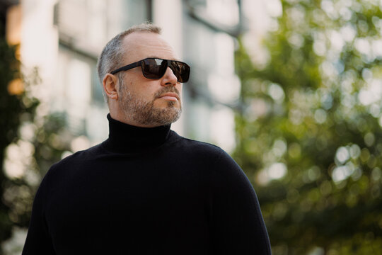 Confident adult man in sunglasses and black turtleneck standing on city street