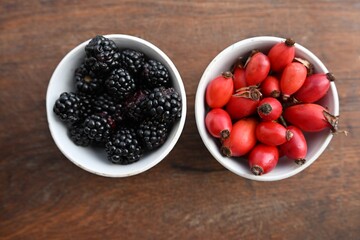 Flat lay of wild blackberries and red rose hips in two white bowls. Highlighting superfoods, color contrast, antioxidants, and autumn harvest on wood.