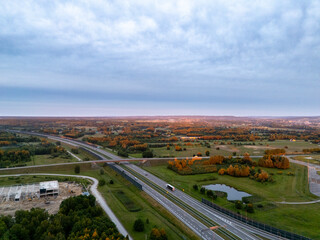 Aerial view of a tranquil landscape during sunset, featuring a winding highway, vibrant autumn trees, and a serene pond, showcasing nature's beauty and traveling routes.