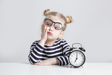 Portrait of Sleepy and Tired Little Girl Wearing Reading Glasses at Table Next to Alarm Clock –...