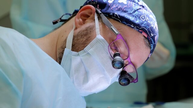 Professional surgeon wearing sterile mask, cap, glasses, surgical magnifier, carefully performing intricate medical operation in pristine operating room environment