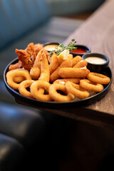 Assorted fried appetizers on a dark plate with dipping sauces, served on a wooden table in a casual restaurant setting. Includes onion rings, mozzarella sticks, chicken tenders, herbs, lemon.