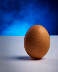A clean and minimalist studio still-life photograph featuring a single brown egg placed upright on a white surface, with a softly lit blue background adding contrast and depth