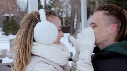 Dreadlocked woman wearing earmuffs playfully covering boyfriend's face with warm gloves during...
