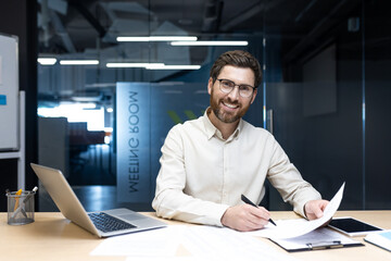 Portrait of a smiling young man working in the office. Sitting at a desk, working on a laptop and with documents