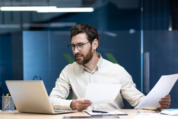 A young man works in an office doing paperwork, holding documents in his hands and looking at the laptop screen