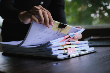 Businessperson organizing and reviewing stacks of paper documents and reports on a wooden desk in an office environment.