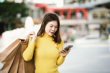 A cheerful young woman in a yellow sweater holds paper shopping bags and a smartphone, enjoying a fun shopping day outdoors.