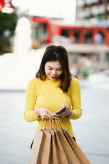 A cheerful young woman in a yellow sweater holds paper shopping bags and a smartphone, enjoying a fun shopping day outdoors.