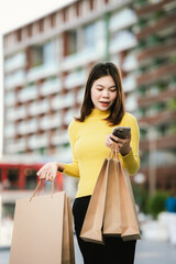 A cheerful young woman in a yellow sweater holds paper shopping bags and a smartphone, enjoying a fun shopping day outdoors.