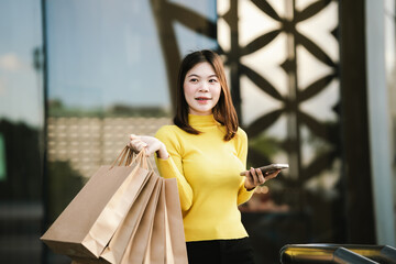 A cheerful young woman in a yellow sweater holds paper shopping bags and a smartphone, enjoying a fun shopping day outdoors.