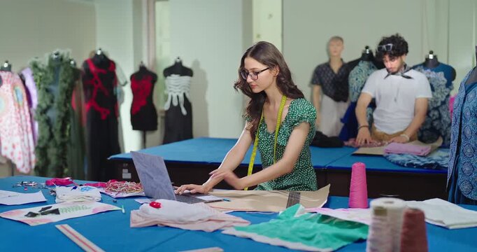 Indian young adult gen z female lady wear tape around neck learn clothe craft skill indoor class room. Pupil girl use laptop look screen hold pencil do fabric pattern on paper new day college place