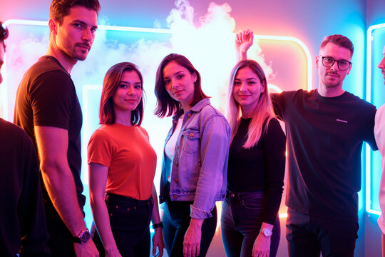 Group of young adults standing together posing confidently in front of neon lights, three women and two men looking at camera with relaxed expressions