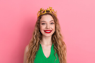 Portrait of a cheerful young woman wearing a golden crown against a pink backdrop and looking directly at the camera © deagreez