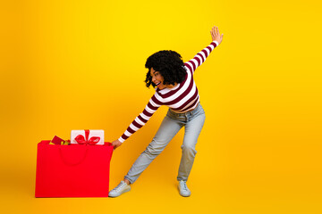 Joyful young woman holding a large red shopping bag with gifts, set against a vibrant yellow background.