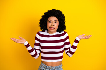 Young woman with curly hair in casual striped top displaying shrugging gesture on bright yellow background