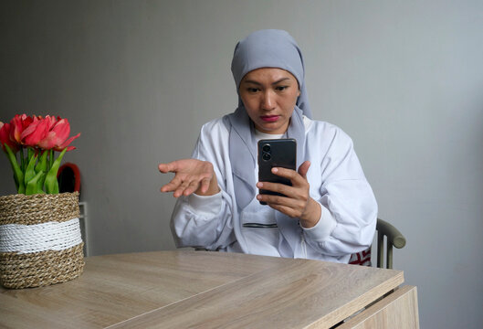 Young Asian Woman In Hijab Talking On The Video Call On Phone While Sitting At Table