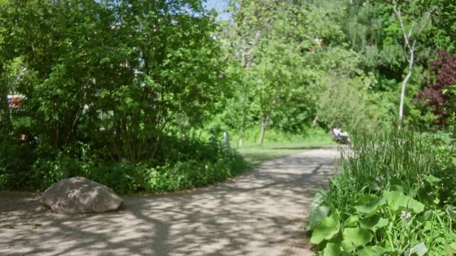 Blurred park path scene with lush green trees and flowers, featuring a defocused figure sitting in the background, creating a tranquil and natural bokeh effect.