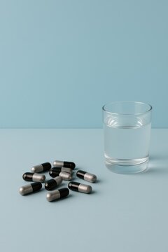 Black and silver supplement capsules beside a glass of water on light blue background, minimalist modern health concept