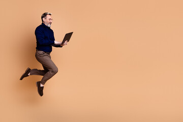 Energetic mature man in formal attire holding a laptop while jumping against beige studio...