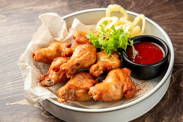 Crispy fried chicken drumsticks with curly fries, red dipping sauce, and fresh herbs on parchment paper over a white plate. Rustic wooden table background, appetizing fast food presentation.