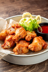 Crispy fried chicken drumsticks with curly fries, red dipping sauce, and fresh herbs on parchment paper over a white plate. Rustic wooden table background, appetizing fast food presentation.