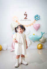 A cute little Asian girl is standing with her birthday party balloons in the background.