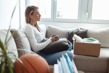Young woman sitting among packed boxes and drinking coffee. Woman drinking coffee while relaxing in...