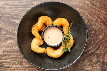 Breaded fried shrimp served on a black plate with dipping sauce and herb garnish, presented on a dark wooden surface. Gourmet appetizer, restaurant-style plating.