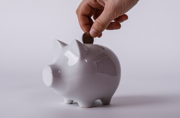 A hand putting a coin into a white piggy bank, representing savings, investment, financial...