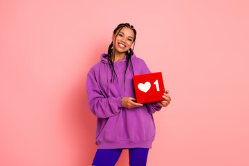 Smiling woman holding a red symbol of a heart, expressing happiness and style, wearing a purple hoodie on a pink background