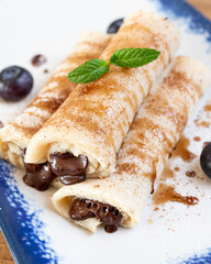 Chocolate filled crepes with syrup, powdered sugar, decorating with mint leaf and blueberries on white plate