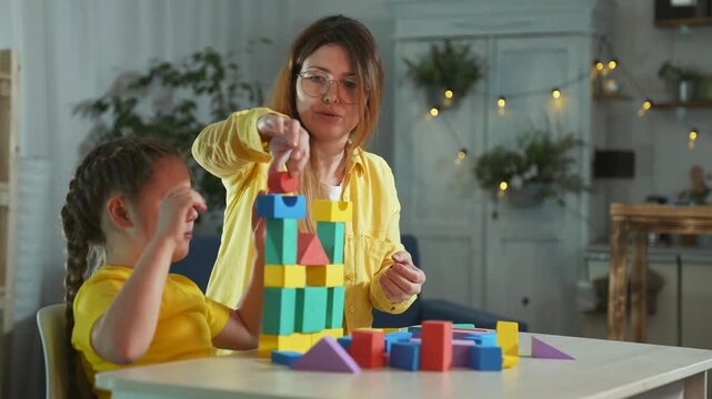 Mother and daughter play with blocks at table in home with colorful wooden toy block tower, parent guiding child through learning, education play fostering development family bonding focus and smile