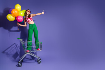Joyful young woman in vibrant attire posing with colorful balloons on a shopping cart against a purple backdrop © deagreez