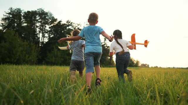 Children run across green field holding wooden toy airplane while kids laugh and play together in tall grass at golden hour capturing carefree playtime energy and friendship during warm summer