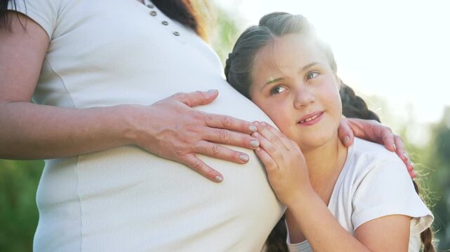 Daughter listening to pregnant belly while resting head against mother hand on belly showing maternity bond family affection outdoors in sunlight with gentle smile protective embrace and love gaze