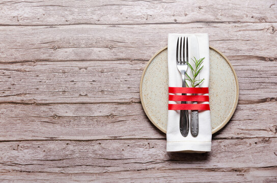 Fork, knife and a napkin tied with a red ribbon and rosemary leaves on a plate on wooden background with copy space.
