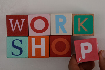 Close-up shot of the word 'WORKSHOP' spelled with colorful wooden blocks, with a hand holding the...