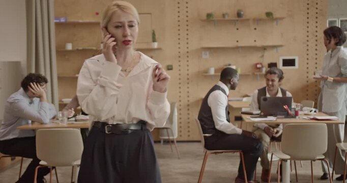 A diverse team works in a busy startup office. A blonde woman talks on her phone, gesturing while other colleagues work and a server brings coffee. - Powered by Adobe